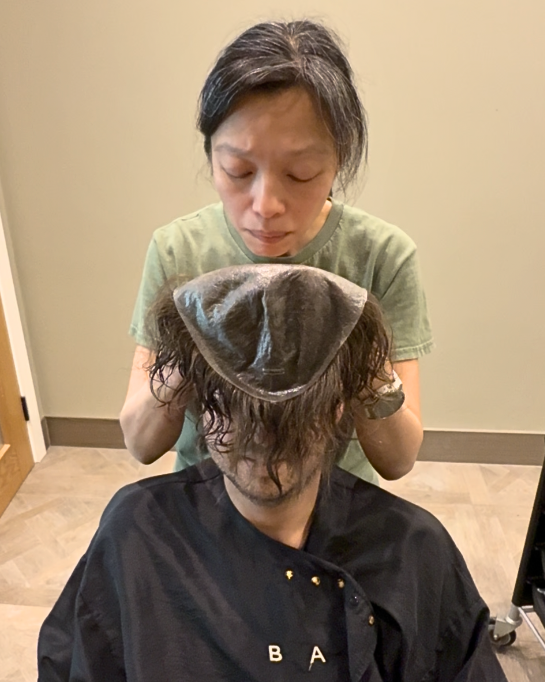 Stylist lowering a hair system base onto a client's head during a fitting