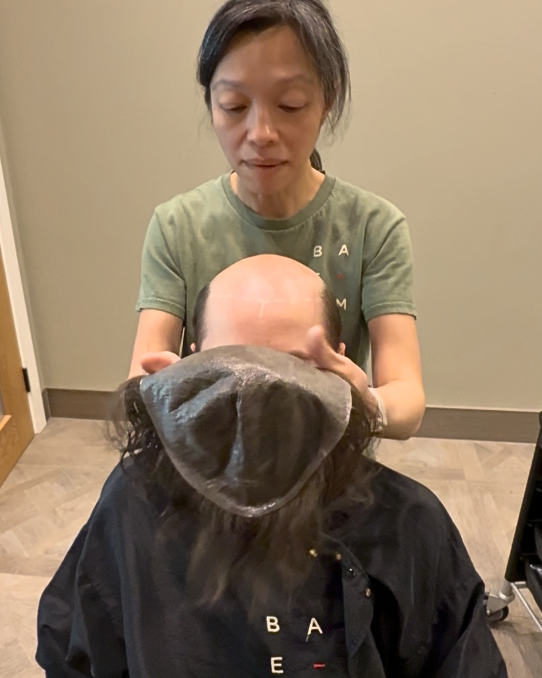 Stylist blending a fitted hair system into a client's natural hairline at a salon mirror